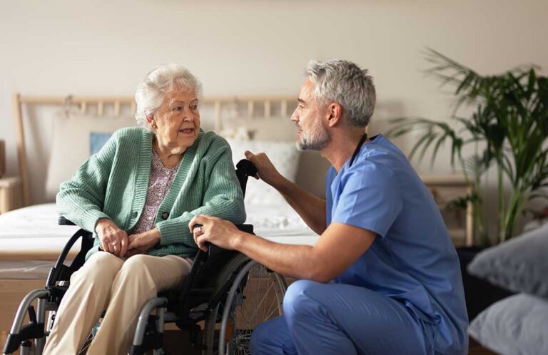An older adult seated at a small table using a tablet device with a gentle smile, natural daylight illuminating the room.